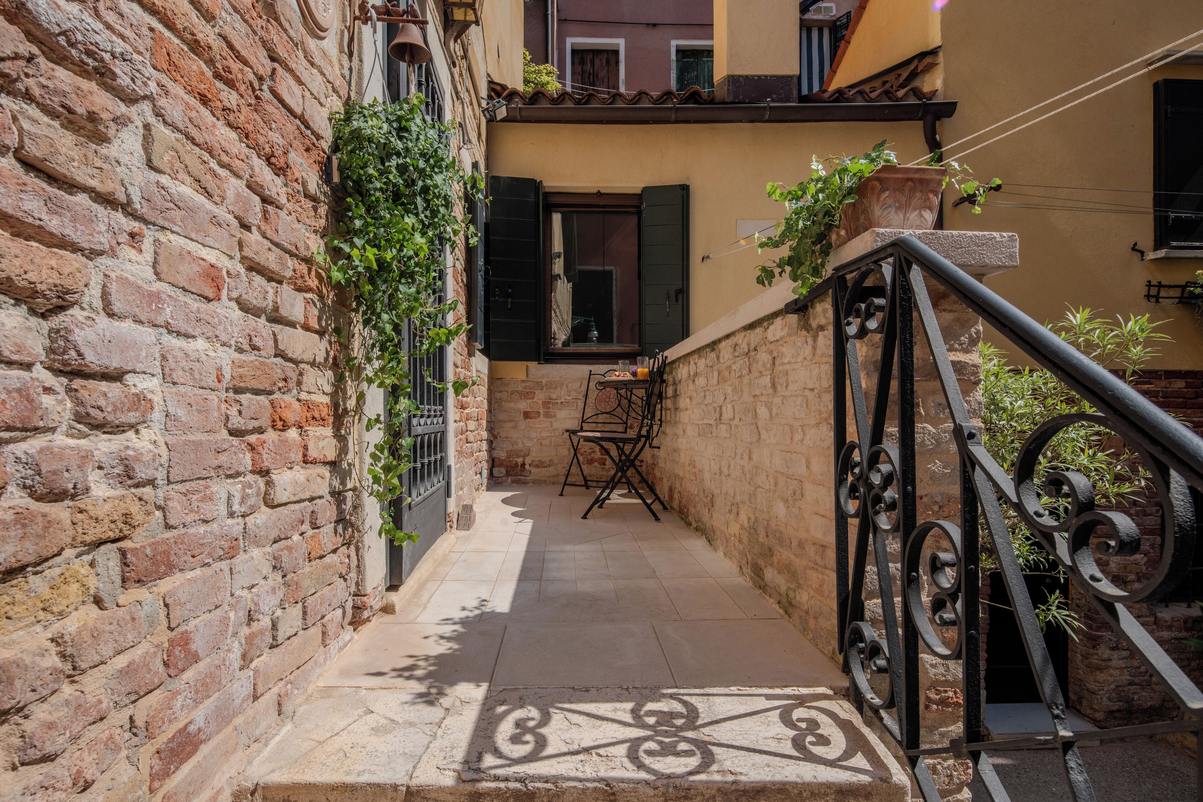 A narrow alleyway with a brick wall on one side and a metal railing on the other. There is a small table and chairs in the center, and a window with shutters.