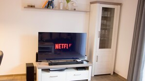 Una sala de estar con un televisor de pantalla plana sobre un mueble blanco, un aparador con puertas de vidrio y un estante sobre el televisor.