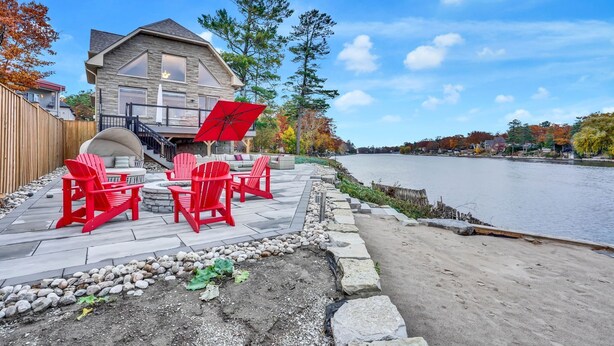A house with a patio overlooking a river, featuring red Adirondack chairs and a red umbrella. - EXECUTIVE WATERFRONT LIVING AT ITS FINEST!!! WELCOME to this immaculate home! (Wasaga Beach)