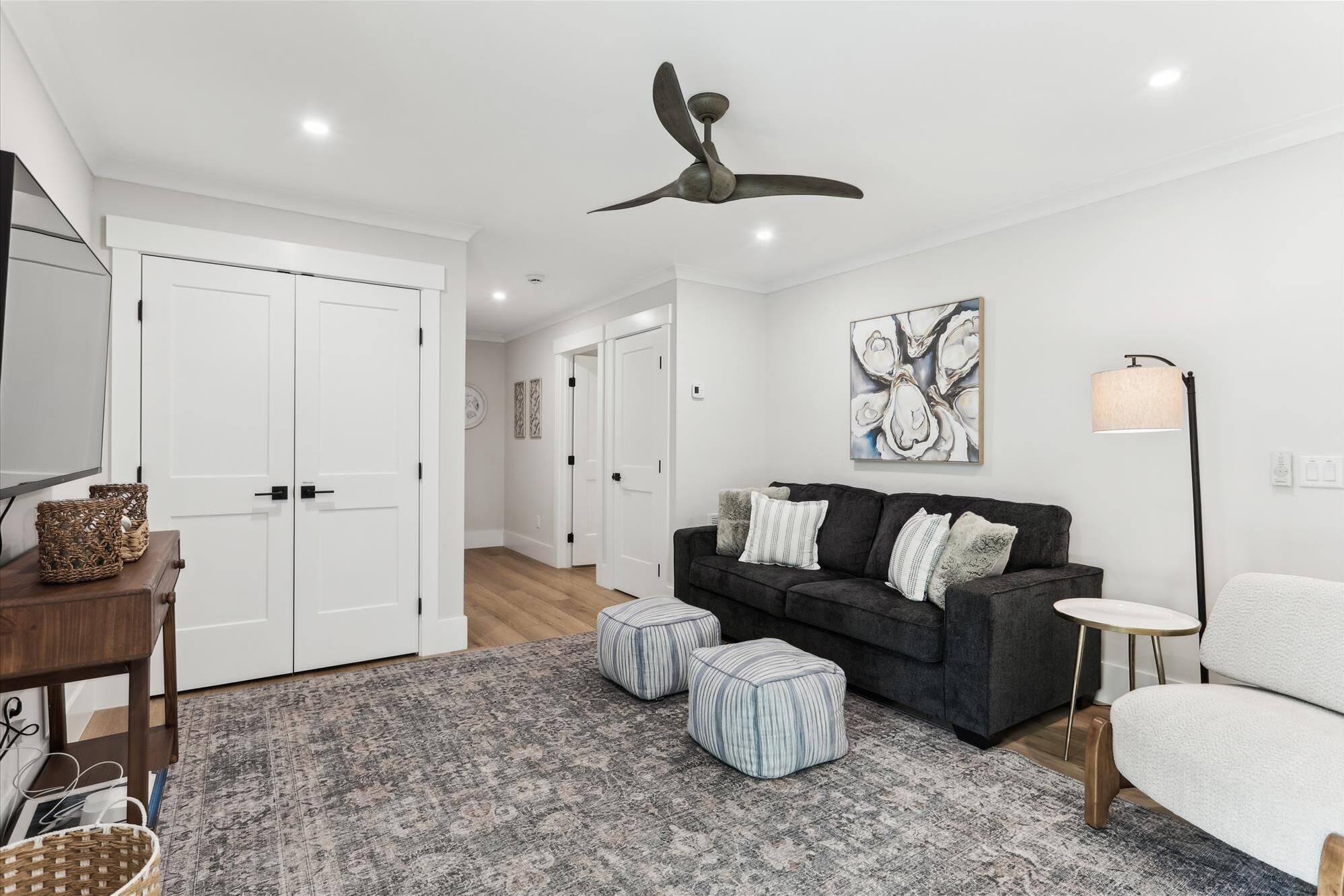 Un salon moderne avec un canapé gris foncé, des poufs à rayures, un fauteuil blanc, une lampe sur pied et un ventilateur de plafond.
