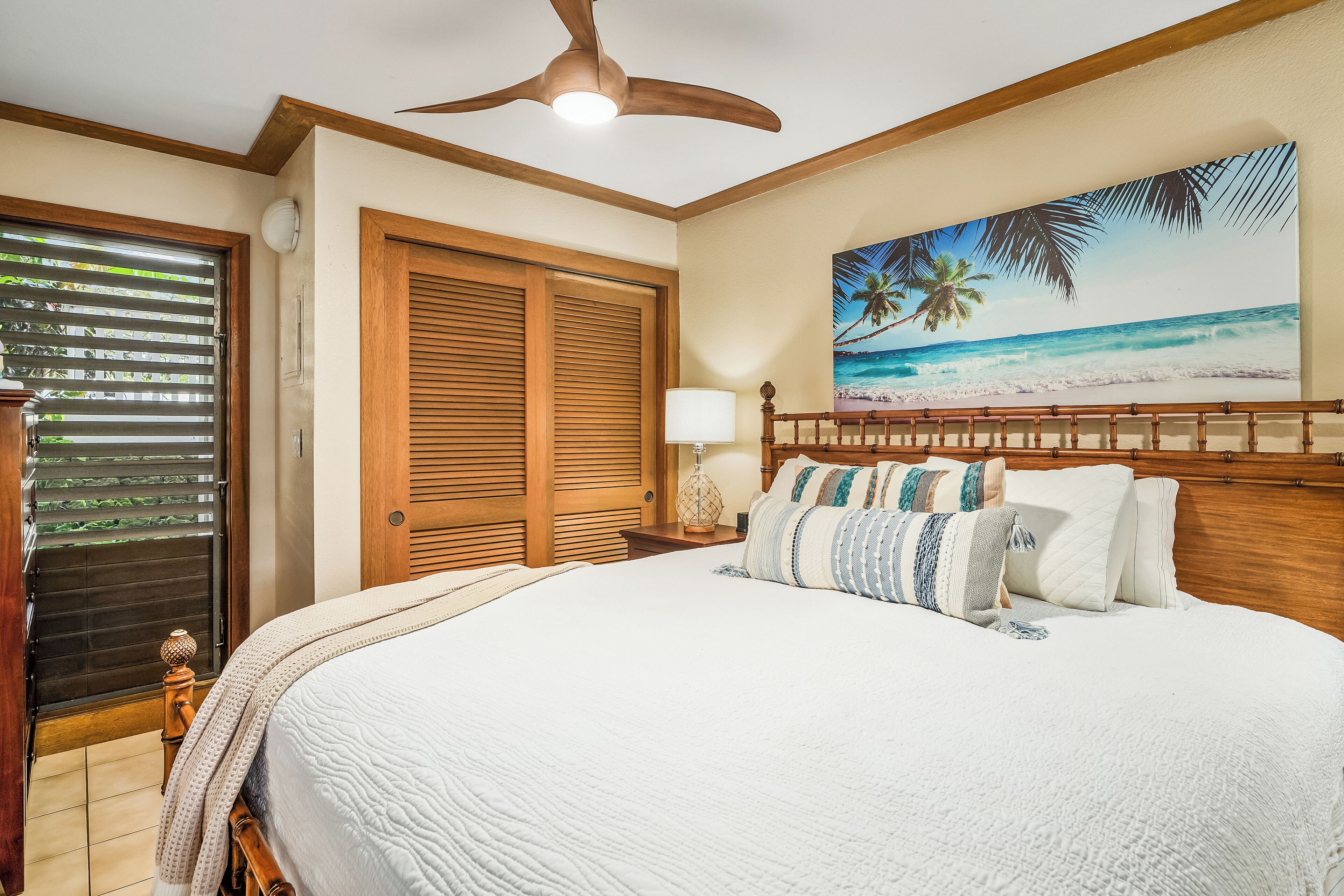 A bedroom with a large bed, wooden headboard, a ceiling fan, a beach-themed painting, and a window with blinds.