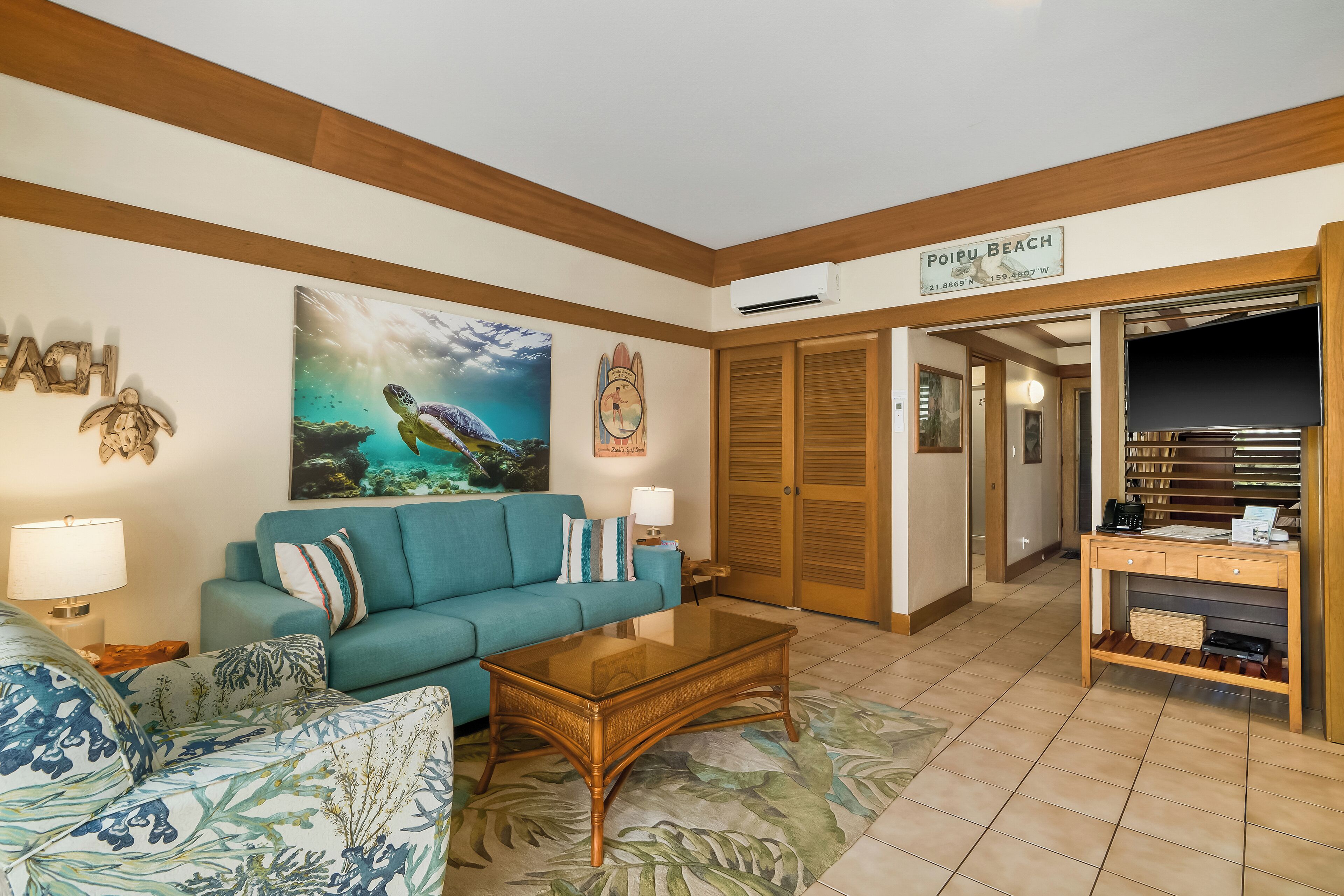 A living room with a blue sofa, a wooden coffee table, and a painting of a turtle. There is a sign on the wall that reads "POIPU BEACH".