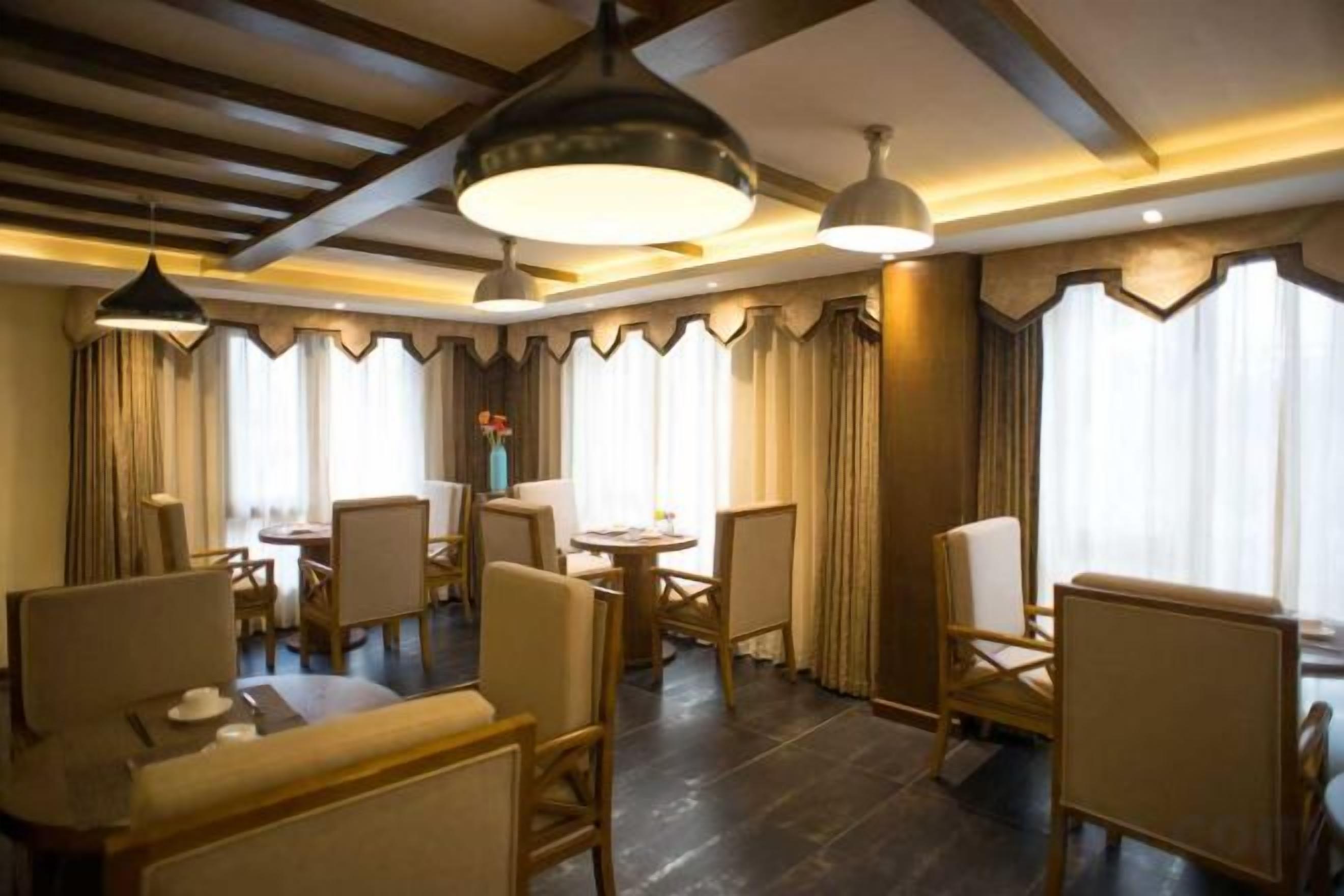 Comedor de hotel con varias mesas y sillas, grandes lámparas colgantes y cortinas.