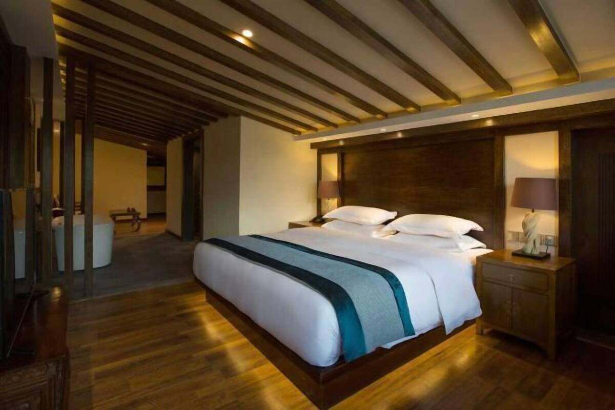 Ein Hotelzimmer mit einem großen Bett, Holzboden, einer Holzbalkendecke, einem Nachttisch mit Lampe und Blick auf den Flur.