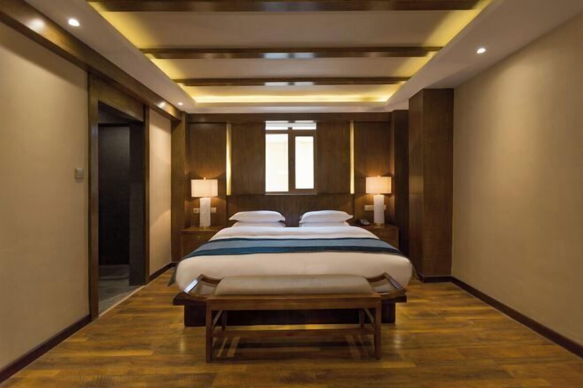 A hotel room with a large bed, two bedside tables with lamps, a bench at the foot of the bed, and a wooden floor.