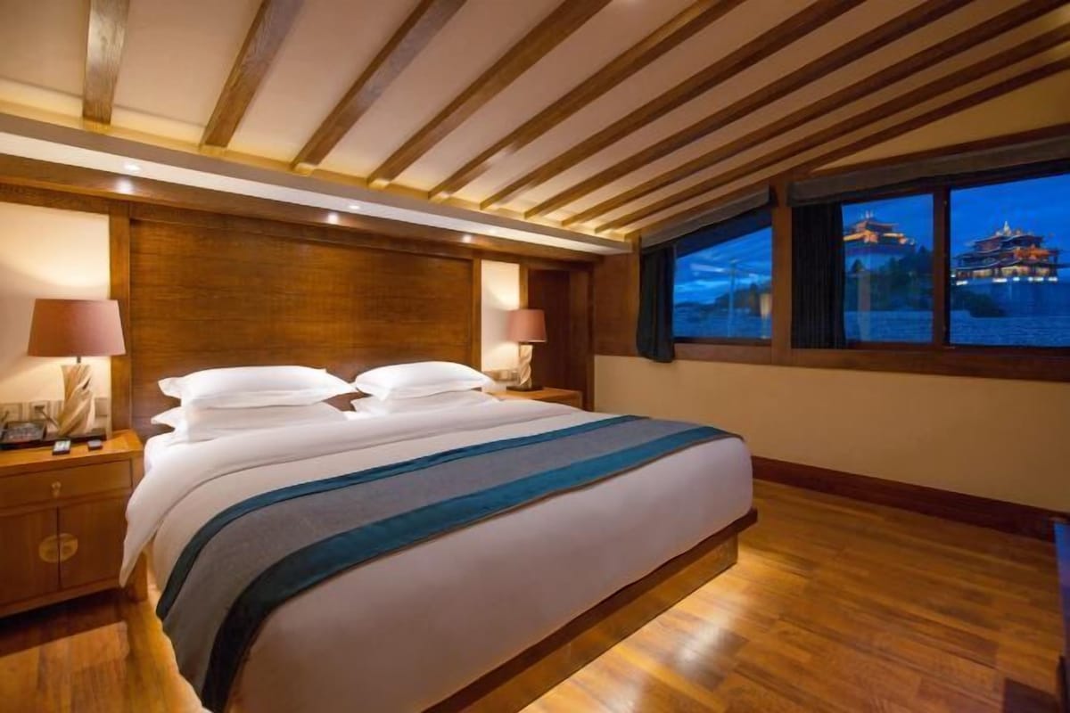 A hotel room with a large bed, wooden ceiling beams, two bedside lamps, and a view of a building across the water.
