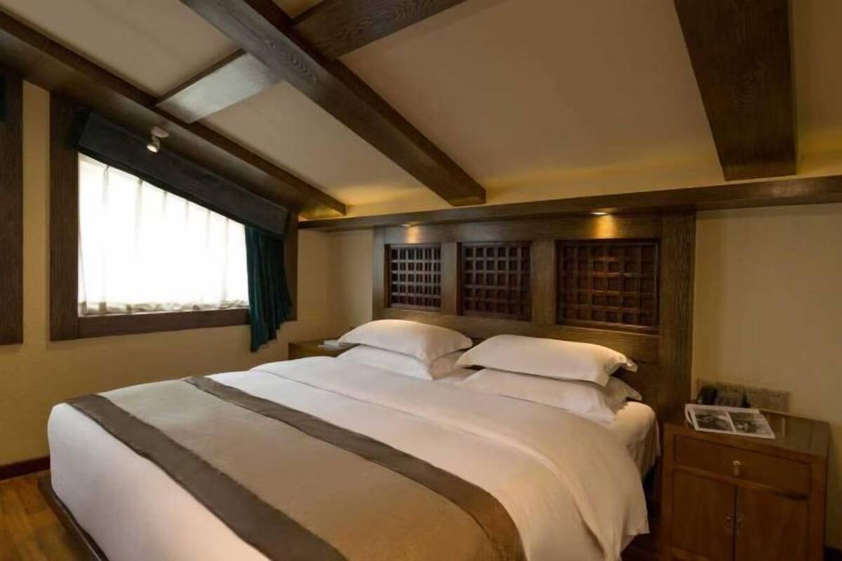 Ein Schlafzimmer mit einem großen Bett, Holzbalken an der Decke, einem Fenster mit Vorhängen und einem Nachttisch mit Lampe.