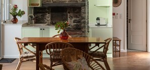 Uma sala de jantar com lareira de pedra, armários verde-claros, cadeiras de vime e uma mesa de madeira.