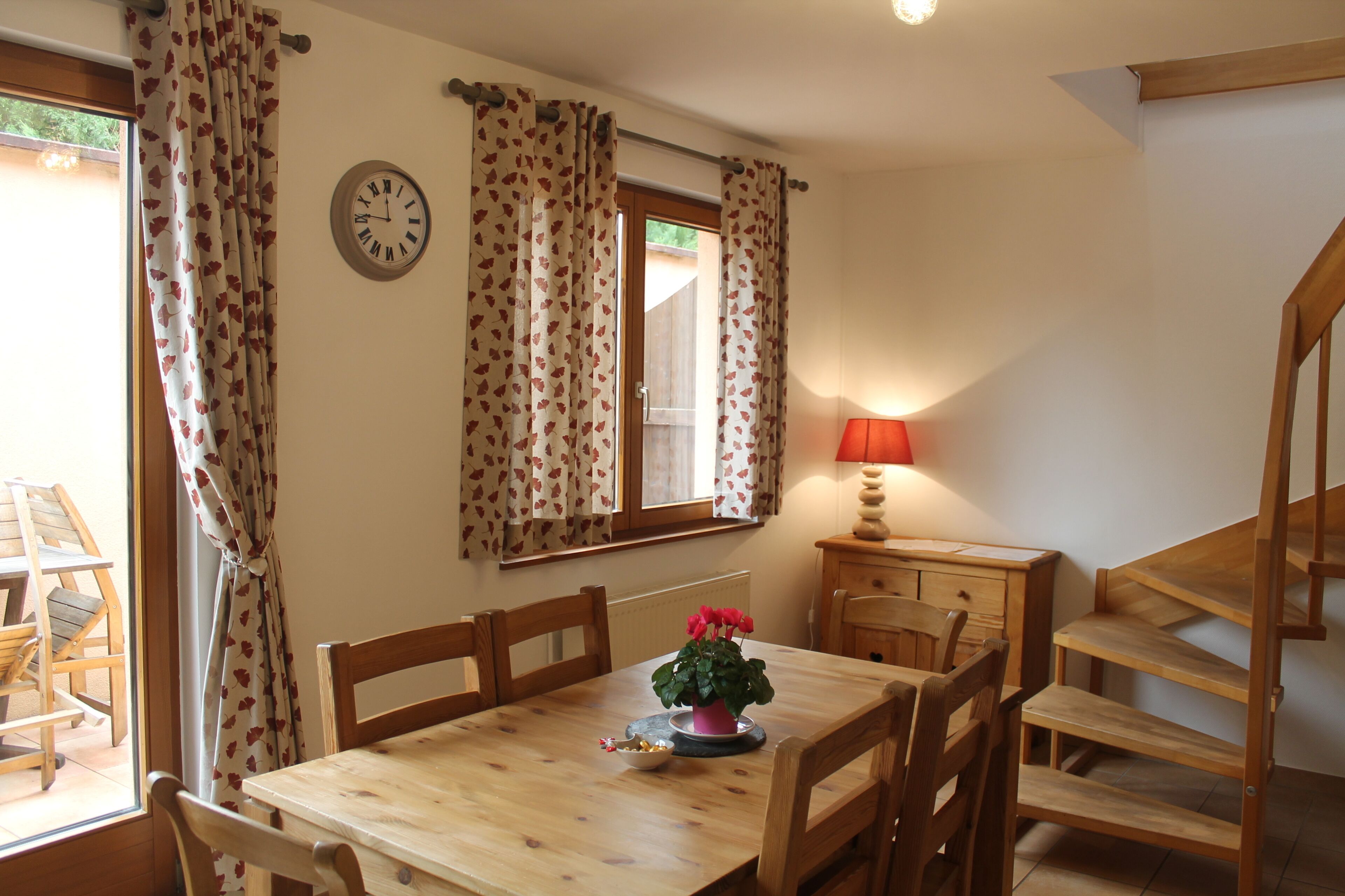 A dining area with a wooden table, chairs, a potted plant, and a clock on the wall.