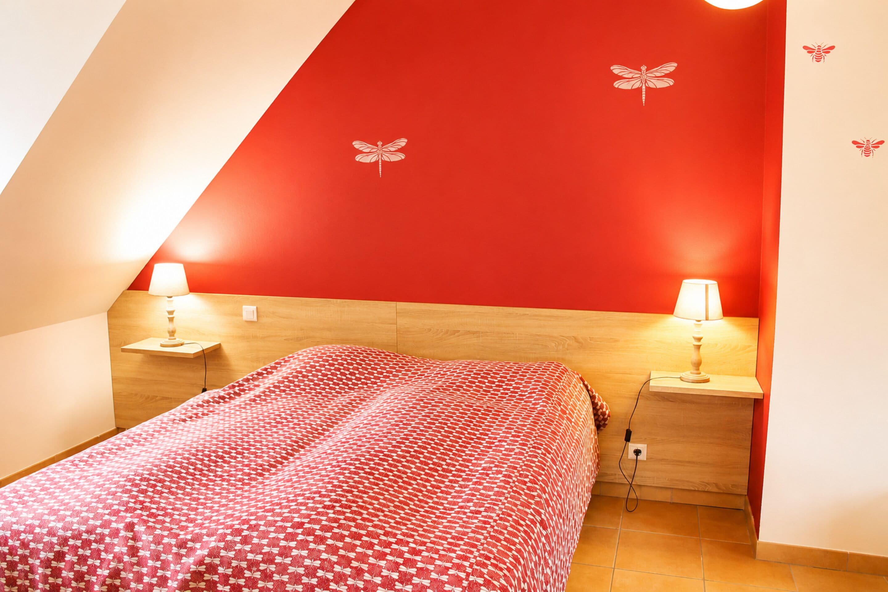 Une chambre avec un mur rouge, un lit recouvert d’une couvre-lit à motifs, deux lampes de chevet et une tête de lit en bois.