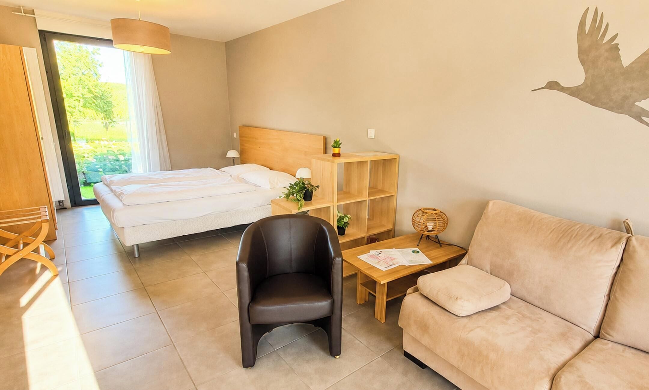Hotelkamer met een bed, een houten nachtkastje, een zwarte stoel, een beige bank en een muurschildering.