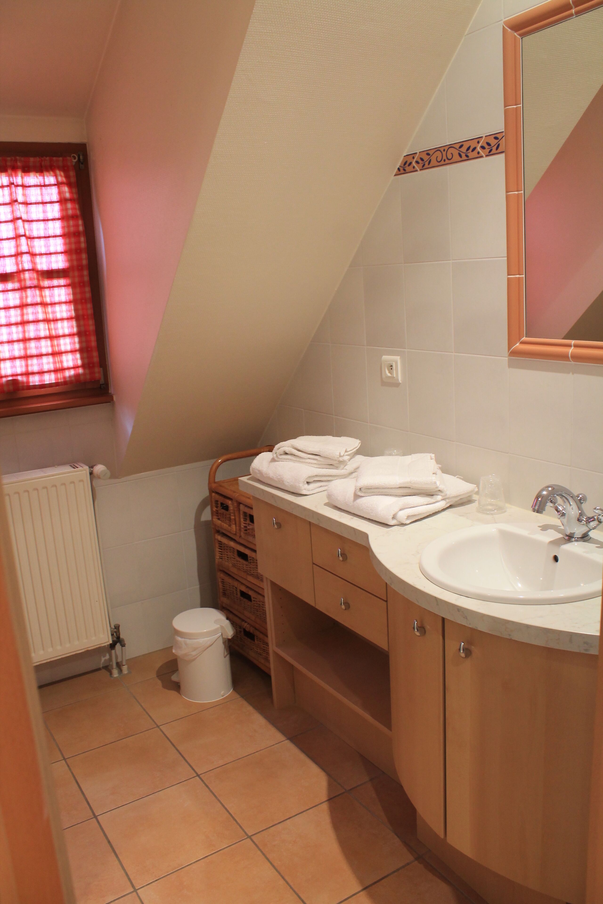 Un baño con un lavabo blanco, armarios de madera, inodoro blanco y una ventana con cortinas rojas.