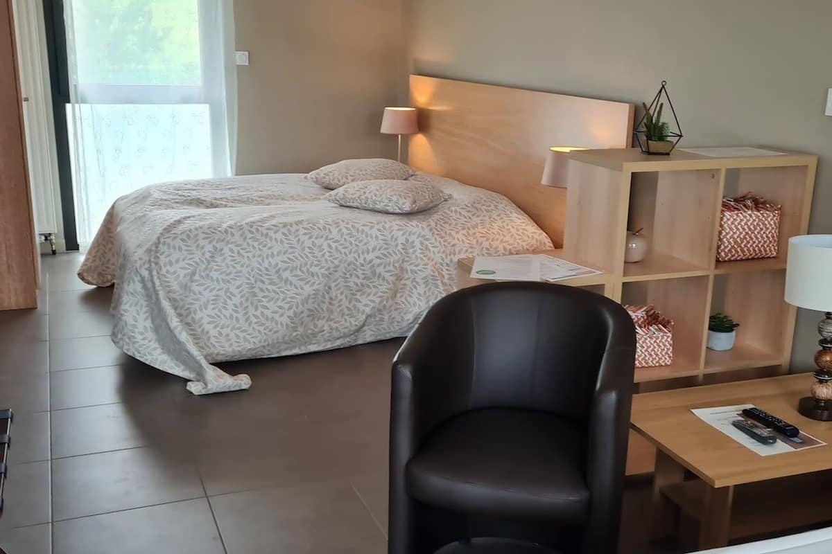 Una camera d'albergo con un letto, una scrivania con una lampada, una sedia e un piccolo ripiano con oggetti decorativi.