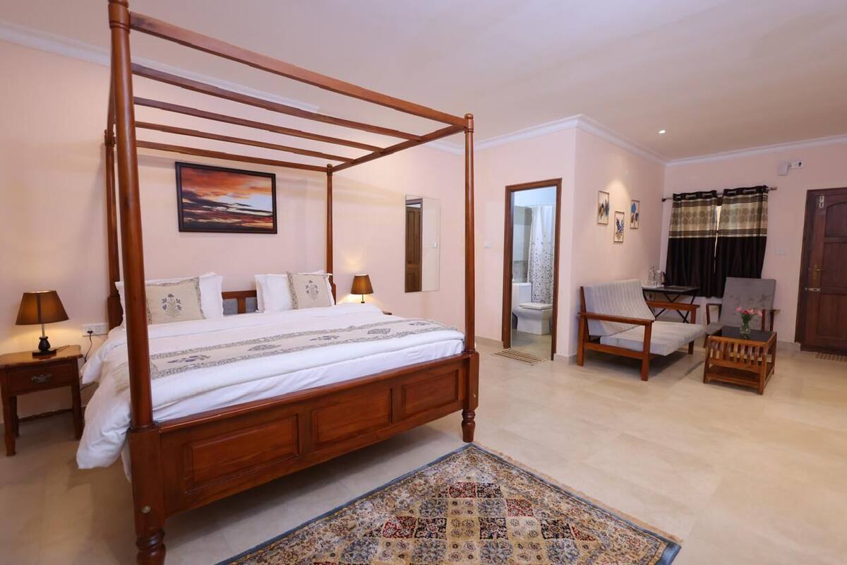 A four-poster bed, a sitting area with a chair and table, a bathroom with a toilet and mirror, and a door leading to another room.