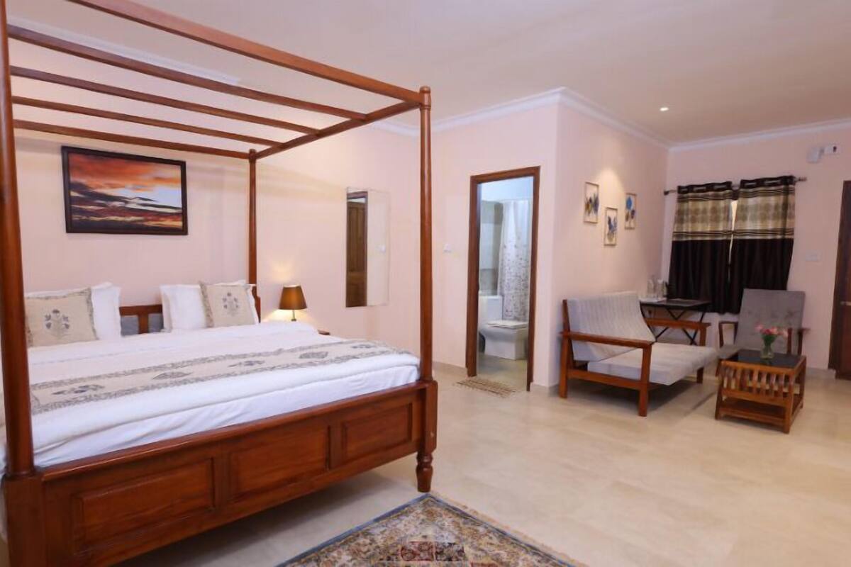 A four-poster bed with a wooden frame, a bedside table with a lamp, a chair, a small table with a vase, and a painting on the wall.