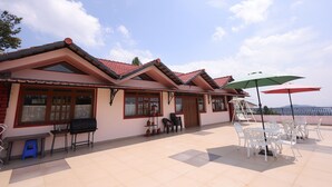 A building with a tiled roof, large windows, outdoor seating with umbrellas, and a view of the sea. - The Forest Haven  (Ooty)