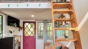 A spacious living area with a loft bedroom above, a wooden ladder, a red door, and a view of greenery outside. - Treehouse with golf cart in Shepherd - near Lake with wild alligators (Shepherd)