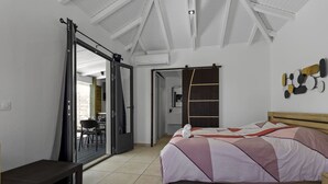 Ein Schlafzimmer mit Bett, einer Bank, einer Tür zu einem Essbereich und einer Wandleuchte.