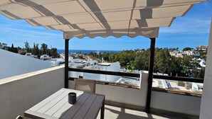 Un balcon avec une table et des chaises, un parasol blanc, et une vue sur la mer et les bâtiments.