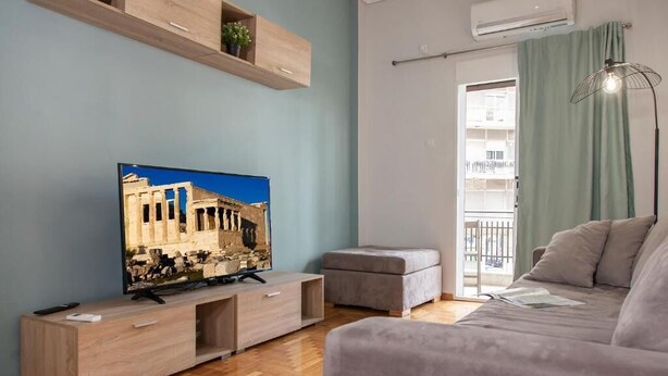 A modern living room with a flat-screen TV, a wooden entertainment unit, a grey sofa, and a balcony with a view. - Mar25 #11 - Entire Apt, Fully Equipped, 1Bedroom. (Athina)