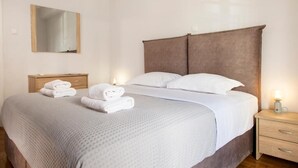 A neatly made bed with white linens and a grey quilt, a wooden nightstand with a lamp, and a mirror on the wall. - Mar25 #11 - Entire Apt, Fully Equipped, 1Bedroom. (Athina)