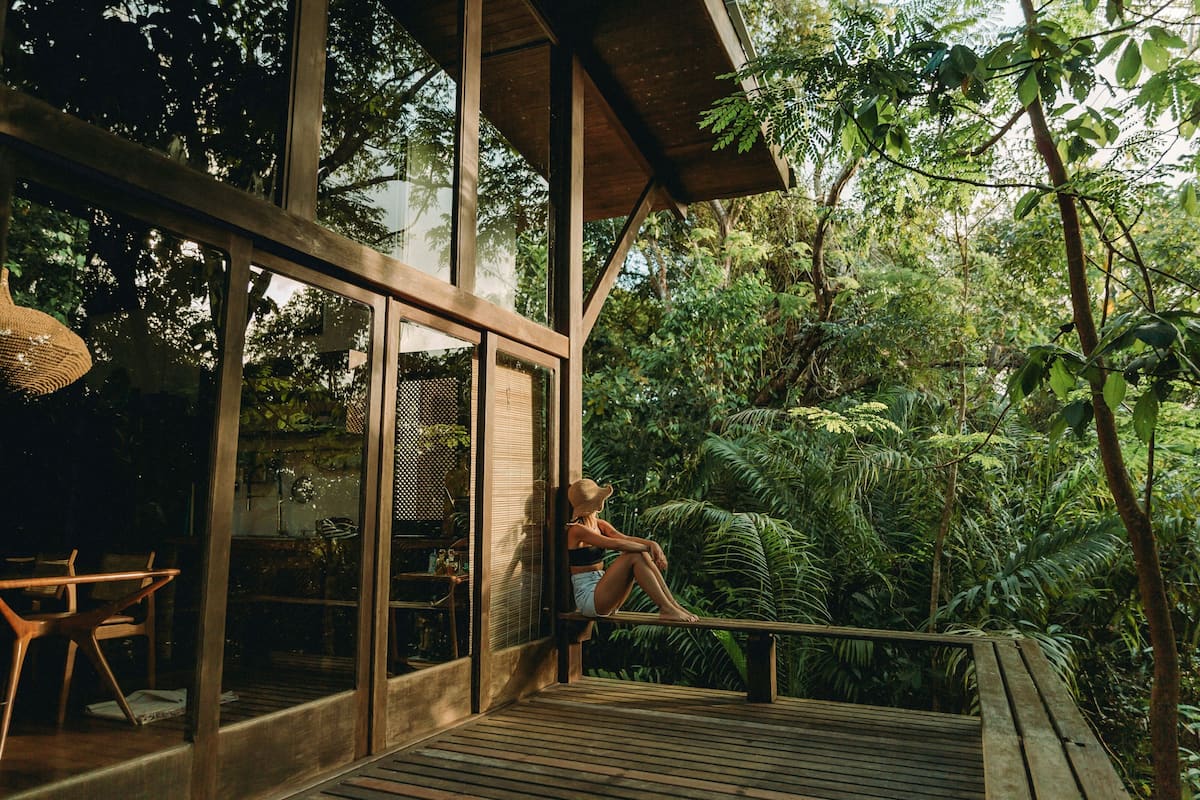 A wooden deck with a person sitting on a bench, overlooking a lush garden with various plants and trees.