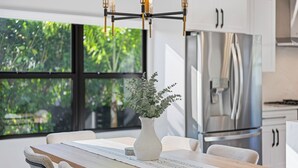 A modern kitchen with a large window, a stainless steel refrigerator, and a wooden dining table with chairs.