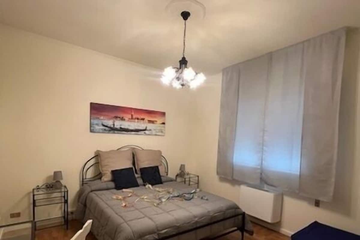 A bedroom with a bed, a chair, a bedside table, a lamp, a painting, a window with curtains, and a ceiling light.