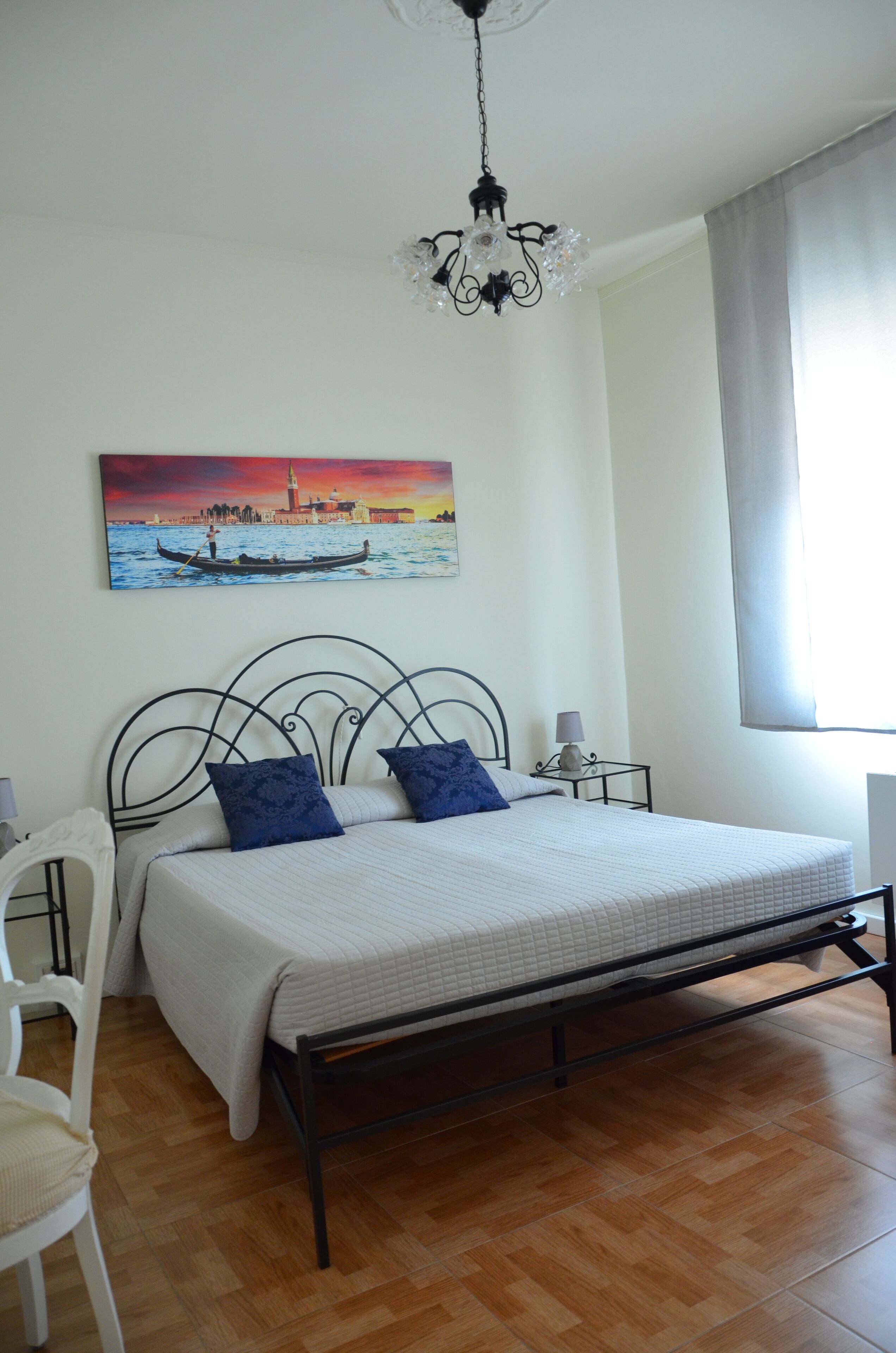 A bedroom with a bed, a chair, a nightstand, a lamp, a painting, and a window with curtains.
