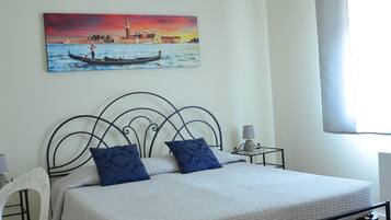 A bedroom with a bed, a chair, a nightstand, a lamp, a painting, and a window with curtains.