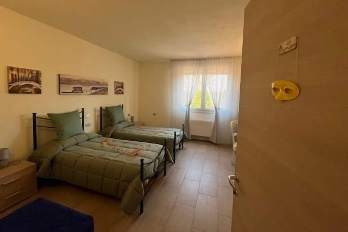 A room with two beds, a small table, and a chair. There are paintings on the wall and a window with curtains.