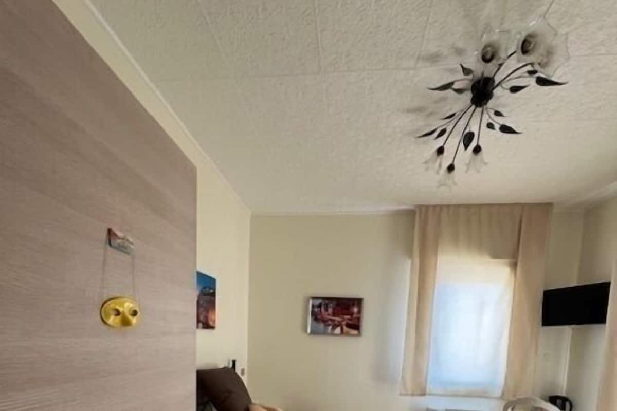 A bedroom with a bed, a desk, a chair, a window with curtains, and a ceiling light fixture.