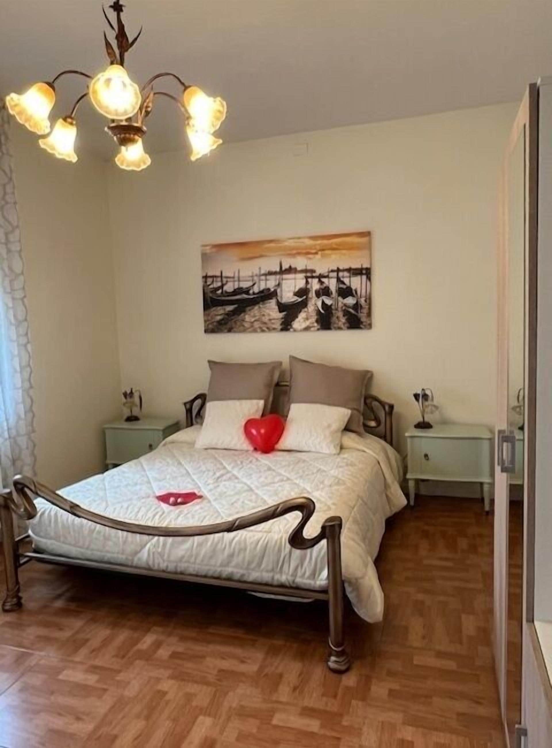 A bedroom with a bed, a nightstand, a bedside table, a painting on the wall, and a chandelier.