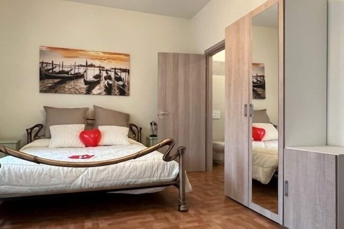 A bedroom with a bed, a wooden wardrobe, a mirror, and a painting on the wall.