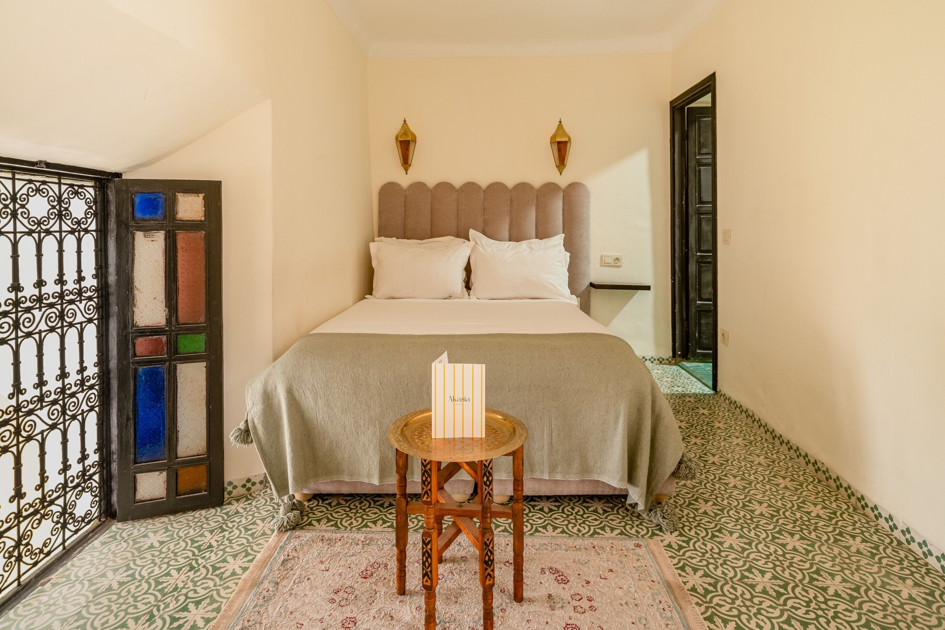 Quarto com cama, uma mesa pequena com uma placa, pisos azulejados estampados e uma porta decorativa.