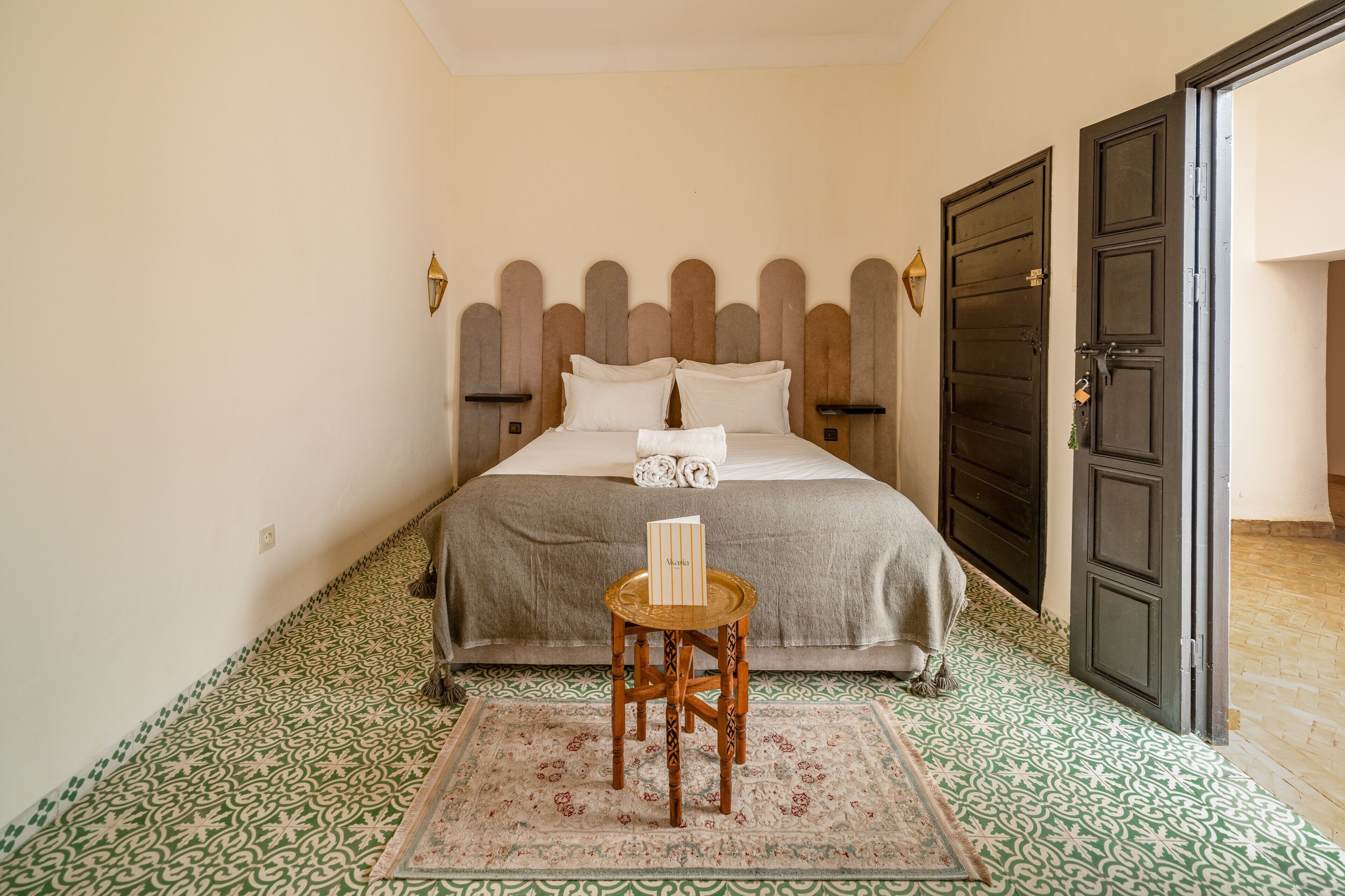 A neatly made bed with a headboard, a small wooden stool with a tray, and a patterned rug.
