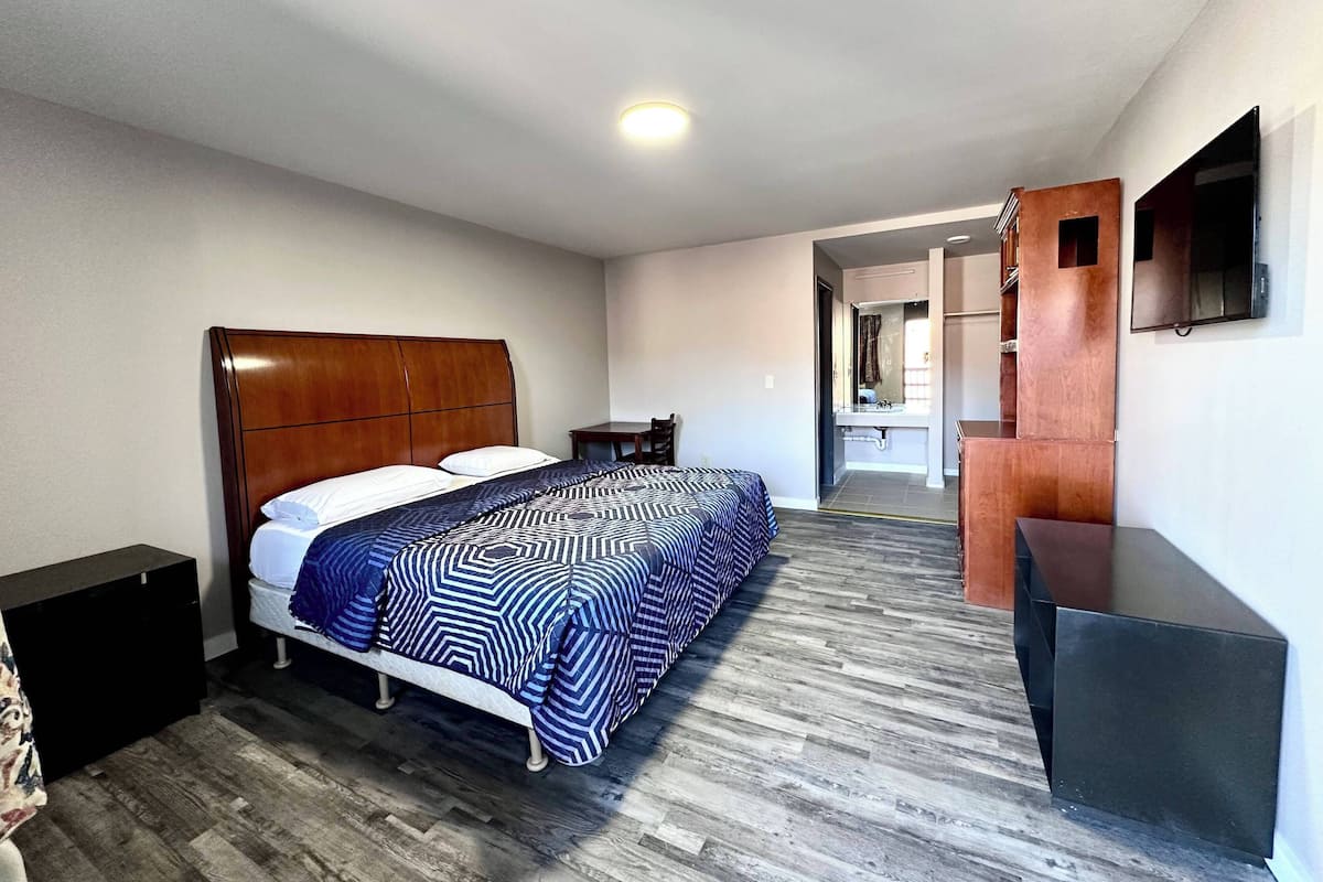 A hotel room with a wooden headboard bed, a patterned bedspread, a small bedside table, a mirror, and a flat-screen TV.