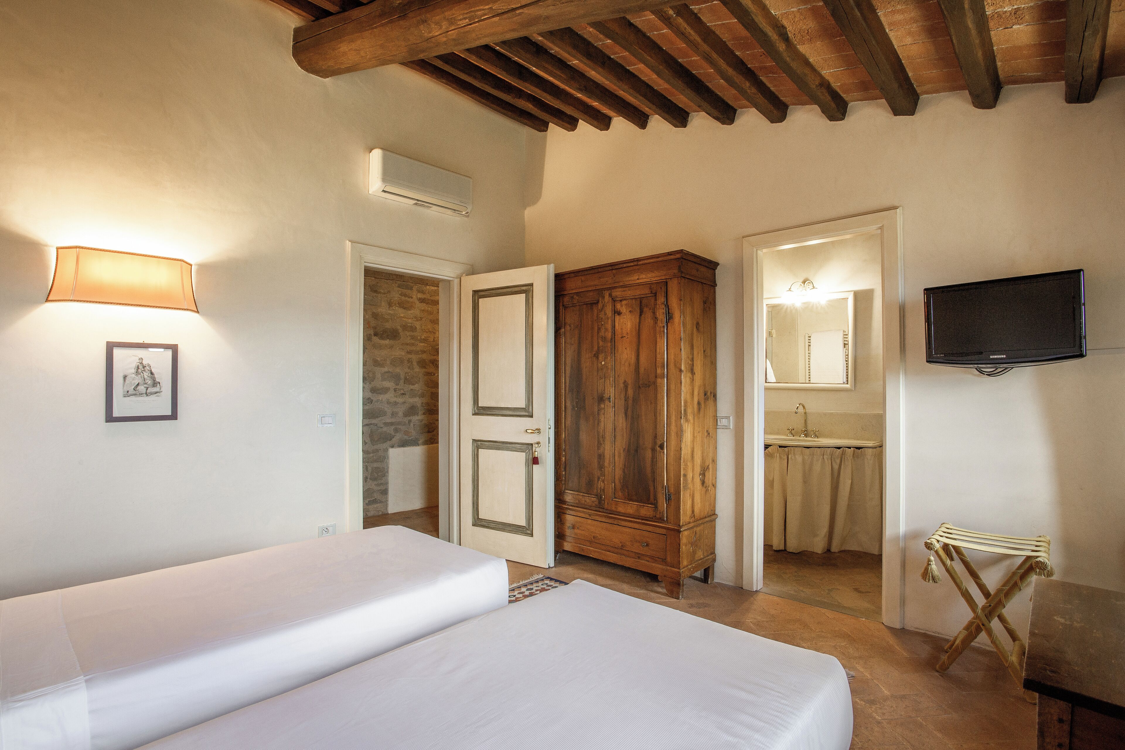 A bedroom with a bed, a wooden wardrobe, a bathroom with a mirror and sink, and a television on the wall.