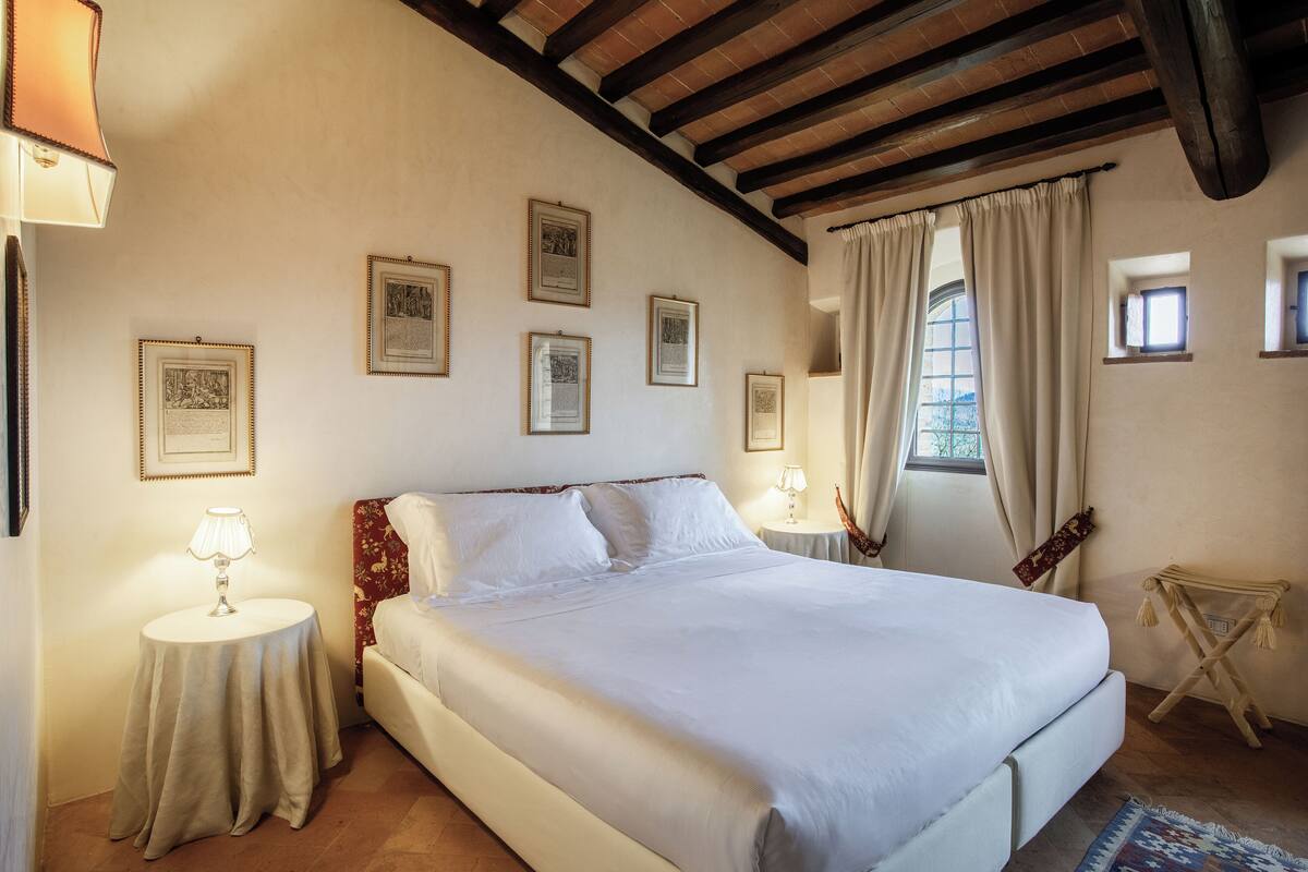 A bedroom with a large bed, a small table with a lamp, and a window with curtains.