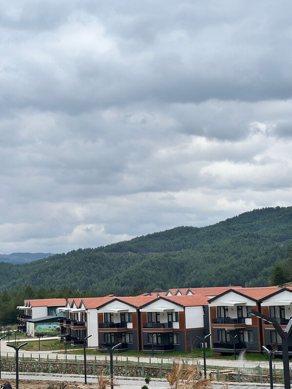 A row of modern, multi-story buildings with red-tiled roofs, set against a backdrop of green hills under a cloudy sky.