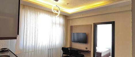 Una habitación de hotel moderna con televisión de pantalla plana montada en la pared, un escritorio pequeño con una silla, un sofá y un ventanal con cortinas.