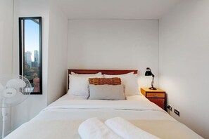 A hotel room with a bed, a nightstand, a lamp, a fan, and a window with a city view.