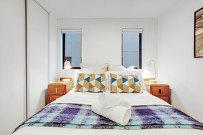 A bedroom with a bed, two bedside tables, a window, and a wall-mounted lamp.