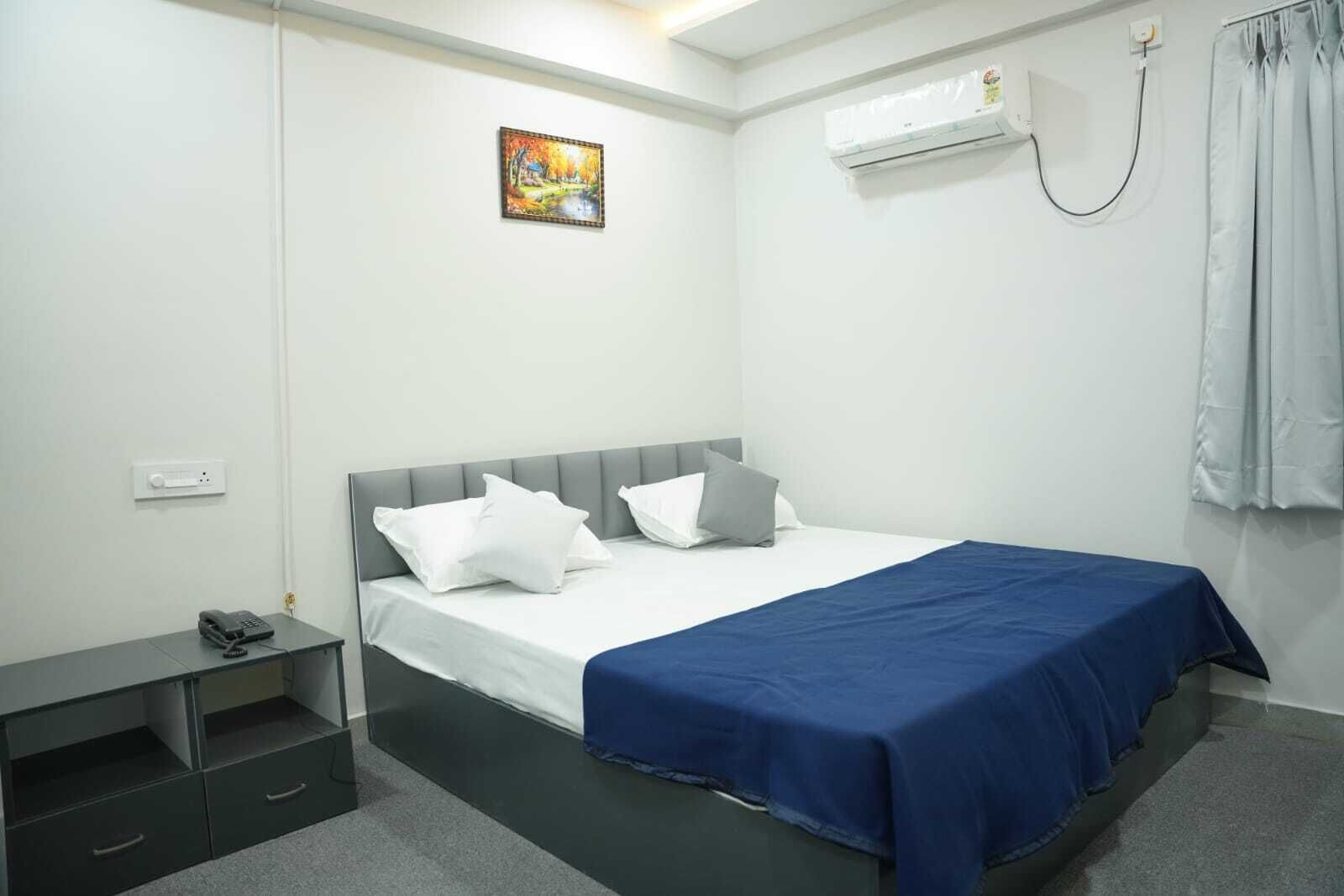 Un dormitorio con cama, mesita de noche, enchufe, teléfono, un cuadro, unidad de aire acondicionado y cortina.