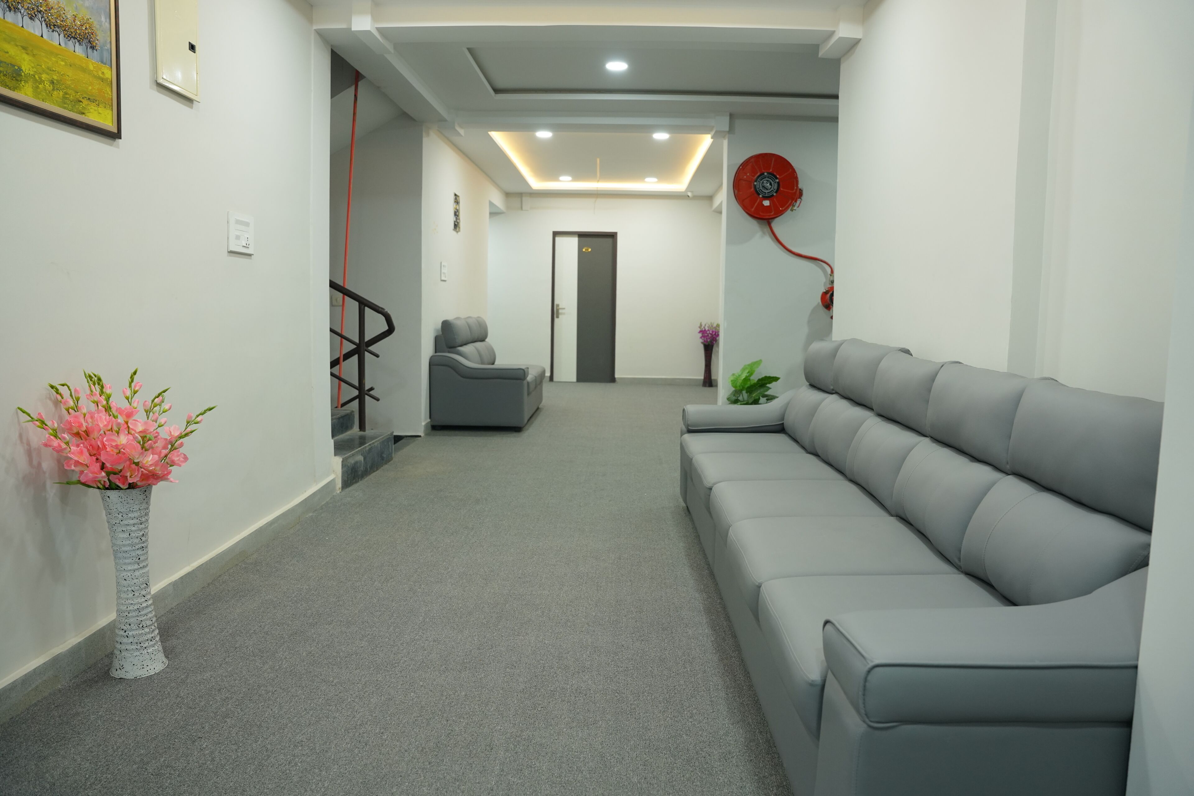 A hallway with a grey sofa, a grey chair, a white vase with pink flowers, and a red wall decoration.