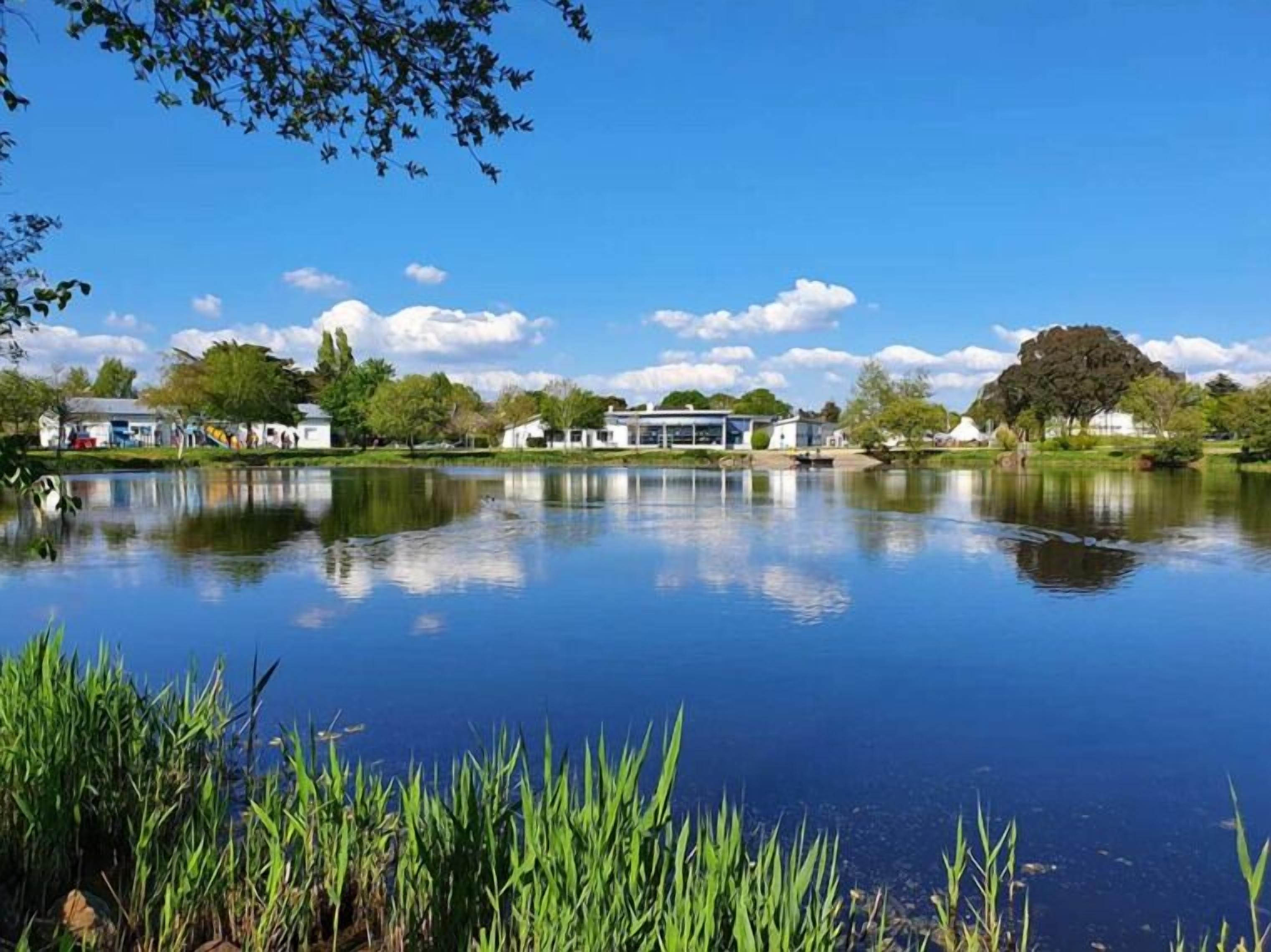 Un lac paisible, dont la surface reflète le ciel et les arbres environnants, et qui est bordé de bâtiments et de verdure.