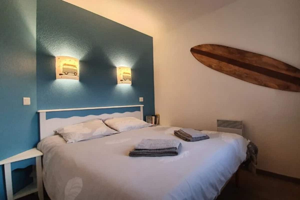 A bedroom with a bed, two wall-mounted lights, a surfboard decoration, and a small cabinet.