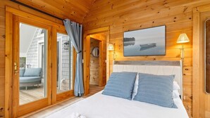 A wooden cabin bedroom with a bed, a window, a door, and a view of the outside. - Quarry Village Compound; 4BR + Hot Tub on a workin (Spruce Head)