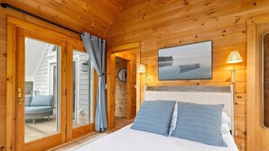 A wooden cabin bedroom with a bed, a view through a glass door, and a painting on the wall. - Pirate's Den on the Wharf (Spruce Head)