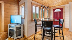 A wooden interior with a TV on a cabinet, a dining table with chairs, and a red door. - Pirate's Den on the Wharf (Spruce Head)